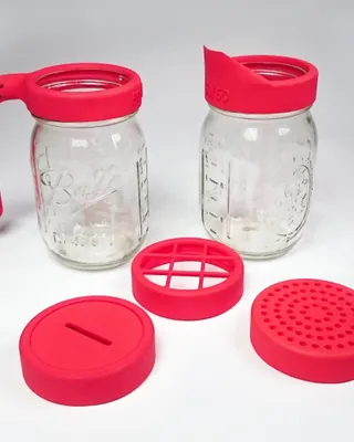 Deux bocaux Mason en verre avec capuchons en silicone rose. Le bouchon gauche a une poignée, le bouchon droit un embout. En face, trois pièces en silicone rose : un couvercle avec une fente, un tamis rond et un autre couvercle perforé.