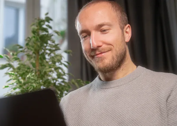 Jeune homme souriant regardant un ordinateur portable dans une pièce éclairée par la lumière naturelle.