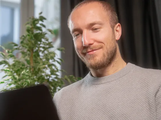Jeune homme avec barbe regardant un ordinateur portable dans une pièce lumineuse avec une plante verte en arrière-plan.