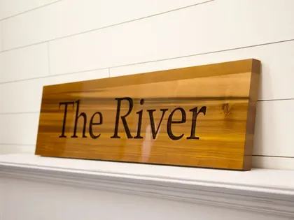 Une plaque en bois gravée portant l'inscription "The River" posée sur une étagère blanche contre un mur blanc avec des lambris horizontaux.