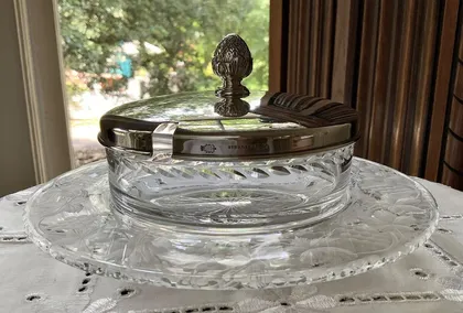 Un plat en verre à motifs, avec un couvercle en métal argenté orné d'une décoration en forme de pomme de pin, placé sur une table avec une nappe blanche.