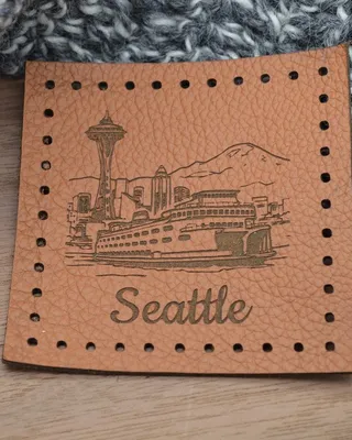 Étiquette en cuir marron avec dessin de la ville de Seattle, incluant la Space Needle et Mount Rainier, et le mot 'Seattle' écrit en bas, posée sur une surface en bois avec un tissu gris en arrière-plan.